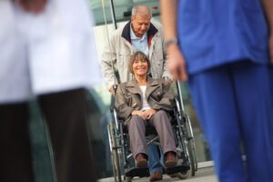 Image of older woman in wheelchair being pushed by husband towards clinicians