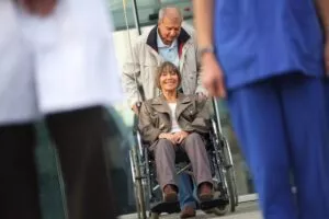 Image of older woman in wheelchair being pushed by husband towards clinicians