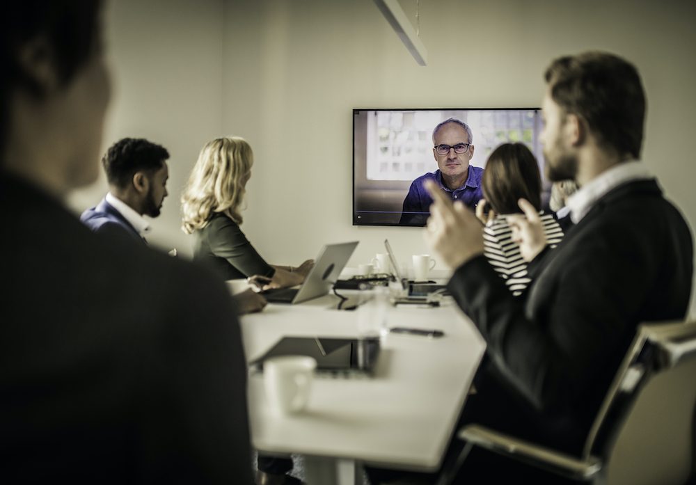 Company board meeting in an office and using a screen to project a manager from overseas during a video conference.