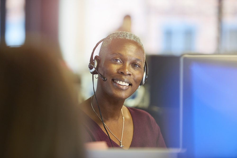 a female customer service representative smiles to camera as she sits at her computer terminal in a call centre chatting happily over her headset to a customer. In the background and foreground colleagues are seen sitting at their desks .