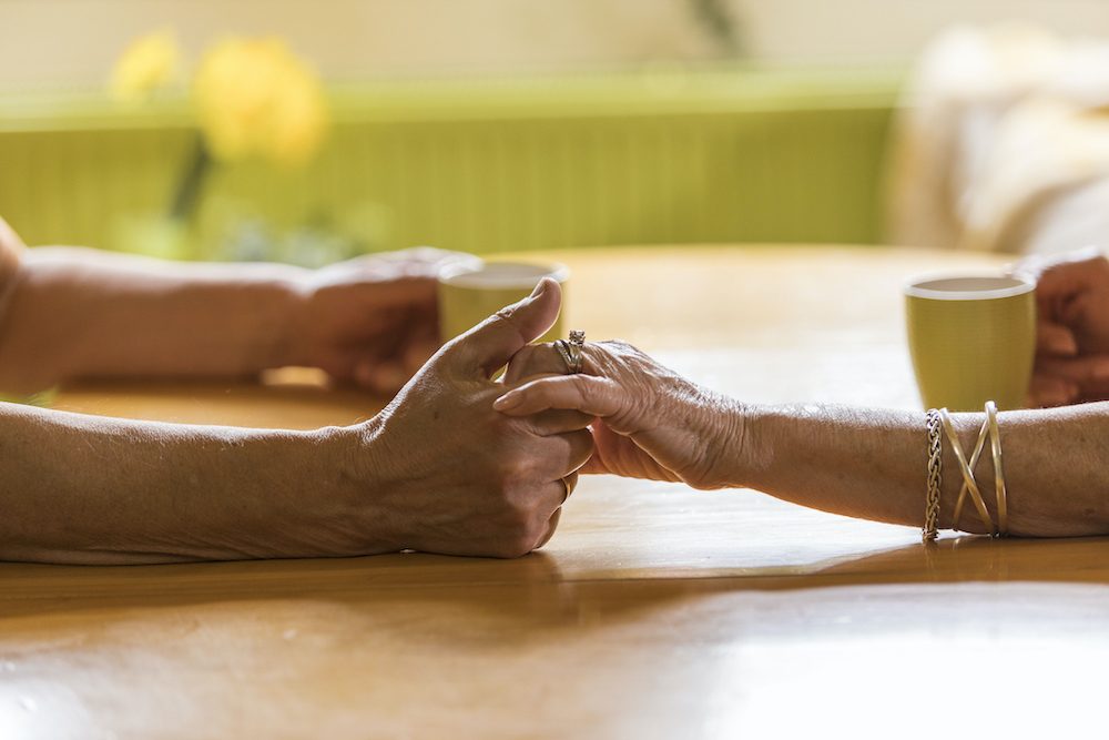 Close up shot of unrecognizable senior couple sitting at table, opposite one another, holding hands while enjoying cups of coffee.